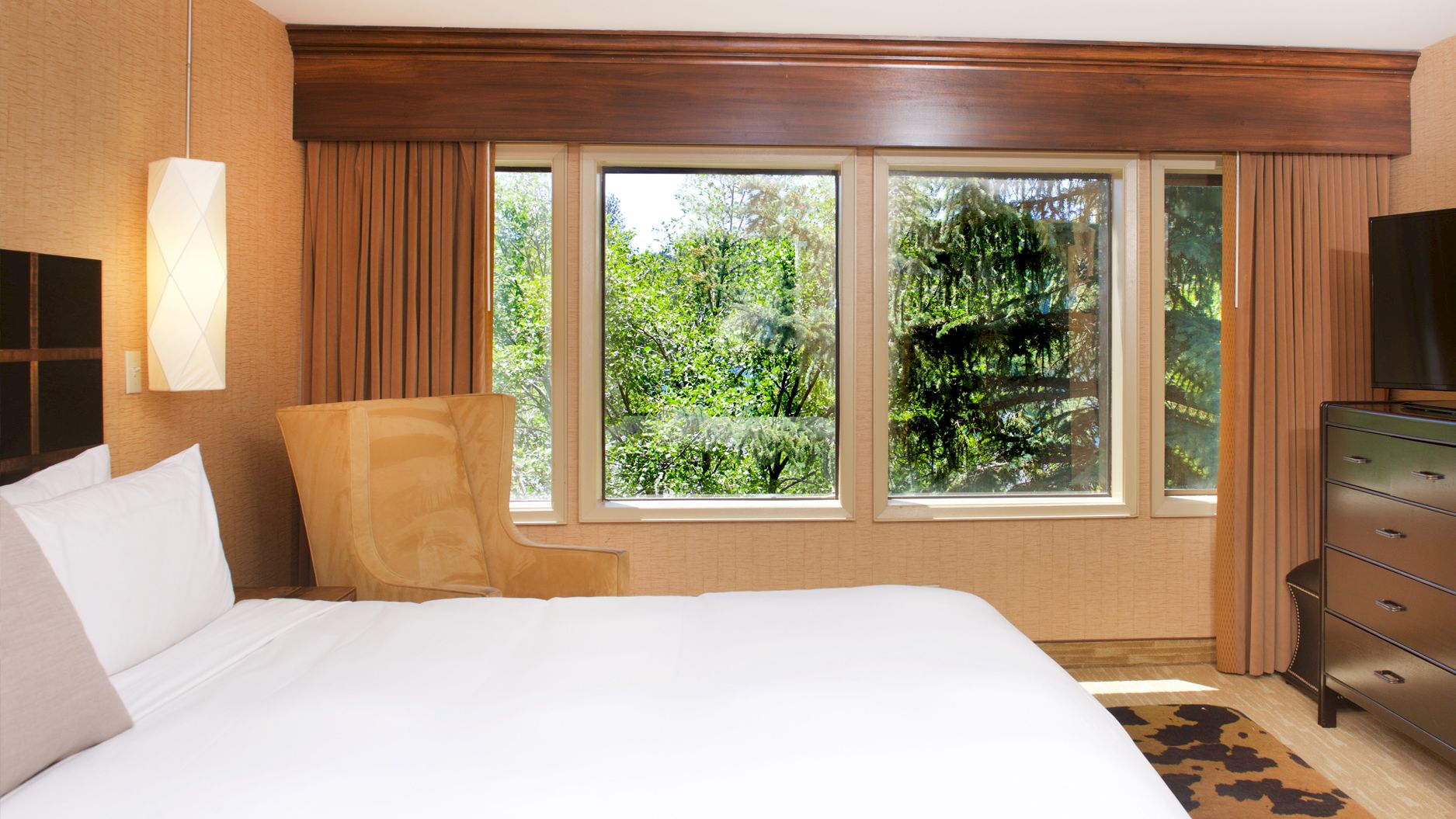 A tidy modern hotel bedroom with a large bed, side lamp, tall windows with curtains, and a dresser.