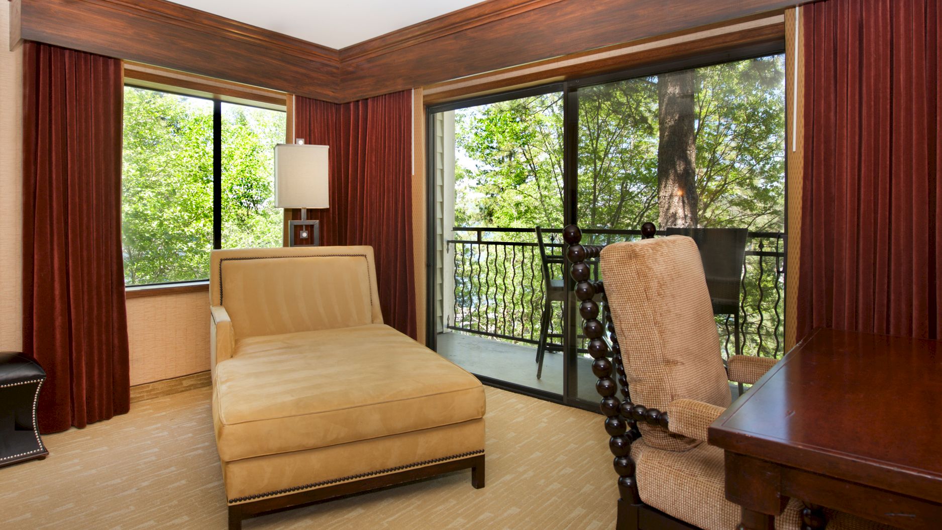 Cozy hotel room with a bed, red curtains, and large glass doors opening to a balcony with lush green trees in view.