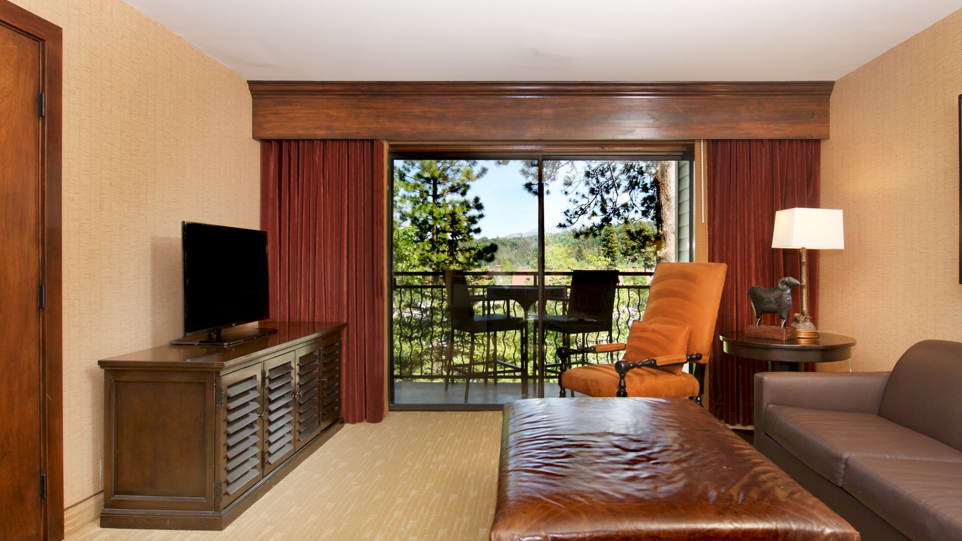 A hotel room with a leather bed, flat-screen TV, curtains, and a balcony view of chairs and greenery, for easy relaxation.