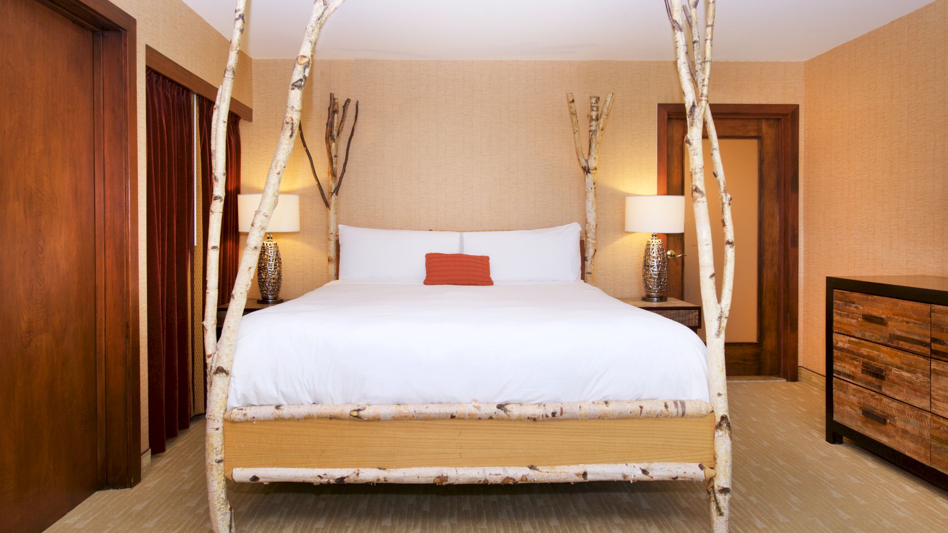 A cozy hotel bedroom with a white bed framed by birch branches, two bedside lamps, and red curtains beside dark wood doors.