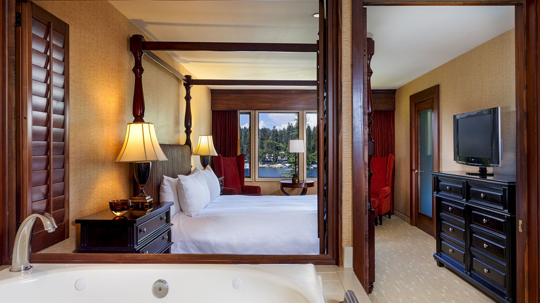 A hotel suite bathroom view looking into a bedroom with a king bed, red curtains, lamps, and a TV on a dresser.