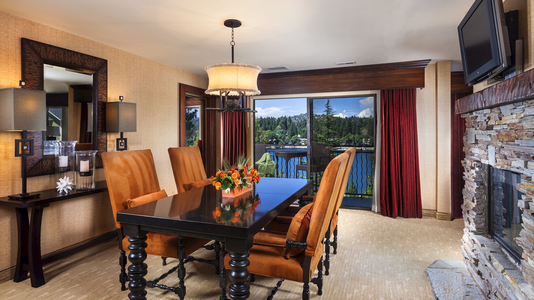 A luxury dining room with a large table, leather chairs, chandelier, red curtains, TV, and a stone fireplace with patio views.