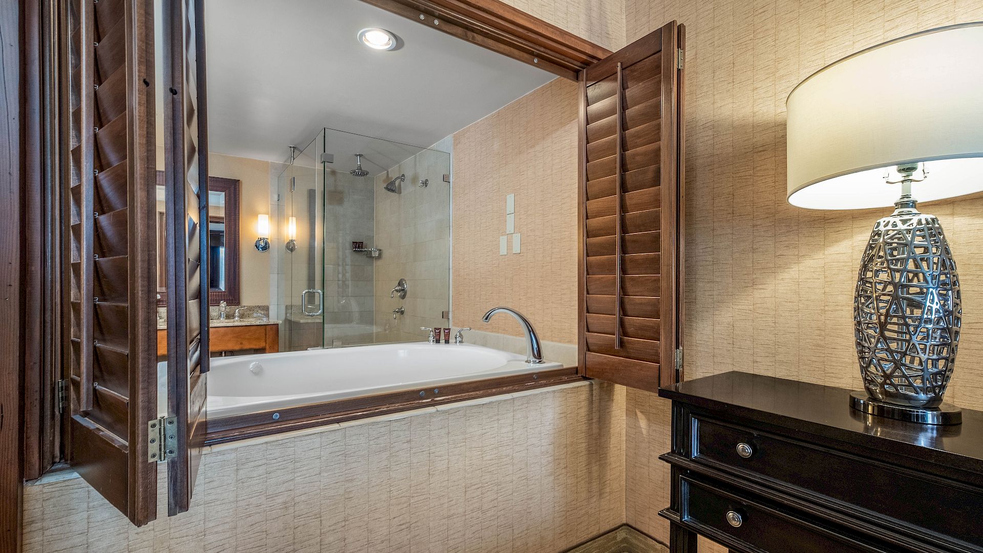 A modern bathroom with a built-in bathtub, glass shower, wood cabinetry, tiled walls, and a decorative lamp on the vanity.