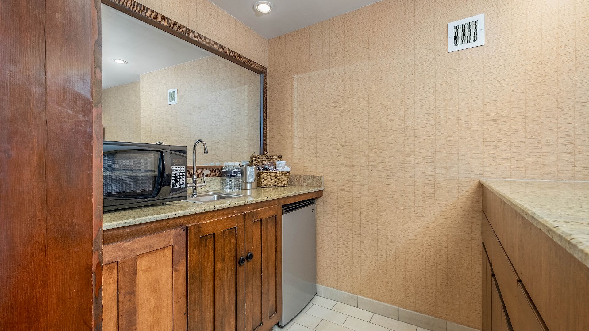 Small kitchenette with a microwave, sink, countertop supplies, wood cabinets, and beige tiled walls.