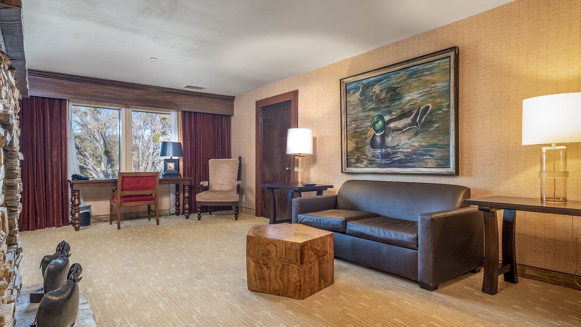 Cozy living room with a blue sofa, wood coffee table, framed duck painting, lamps, chairs, and maroon curtains.