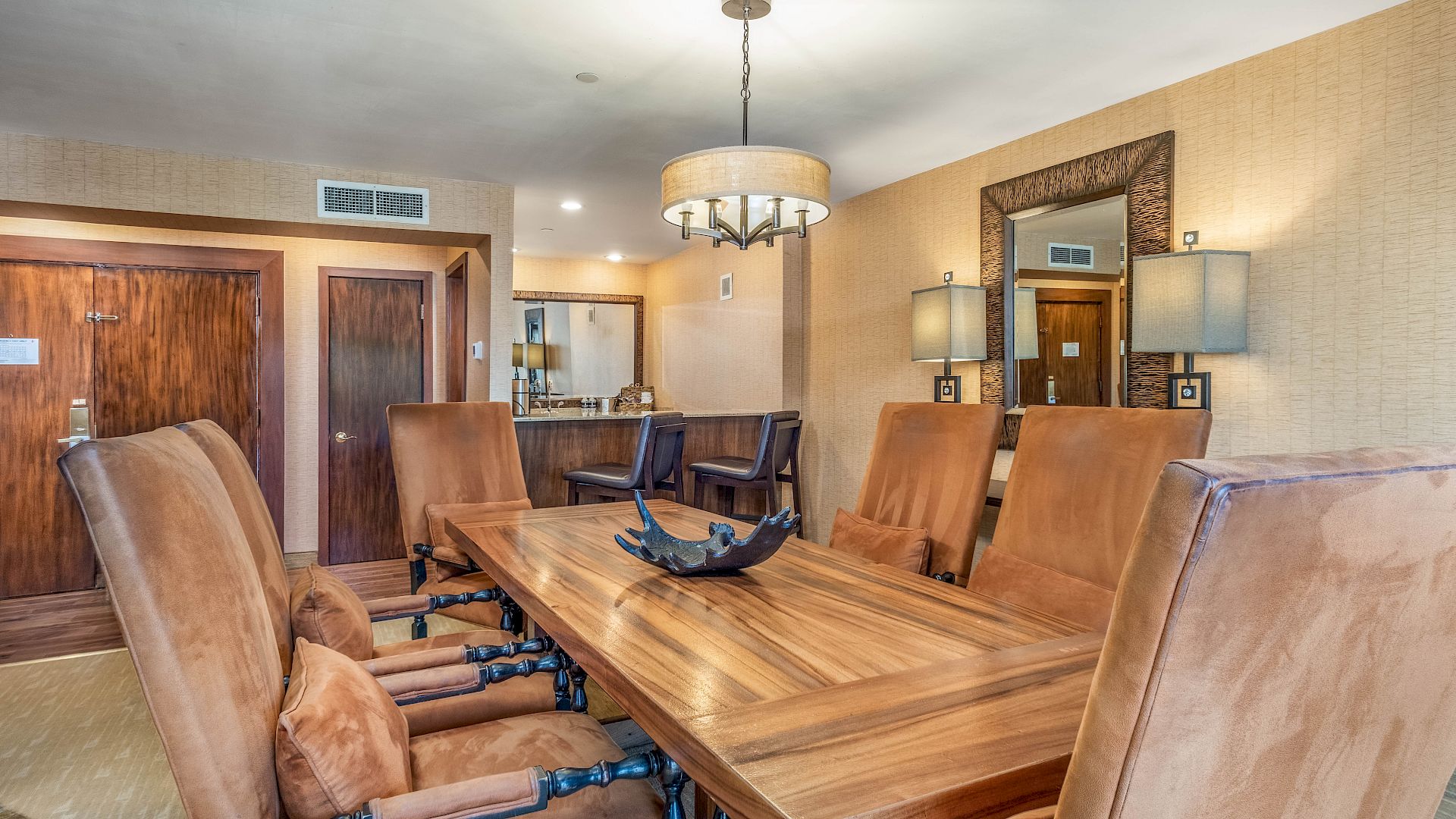 A modern lounge and dining area with a large wooden table, leather chairs, a hanging chandelier, and a kitchen counter in back.