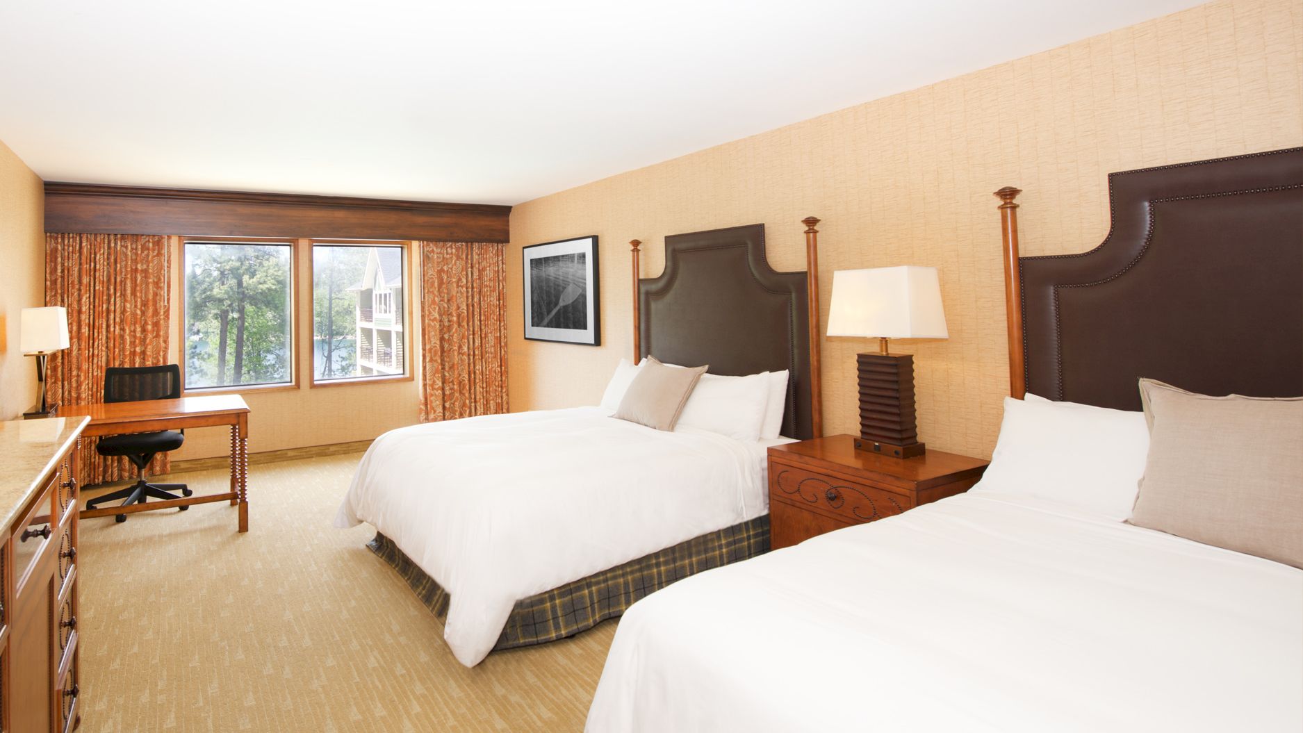 A tidy hotel room with two beds, bedside lamps, patterned curtains, and a desk by the windows.