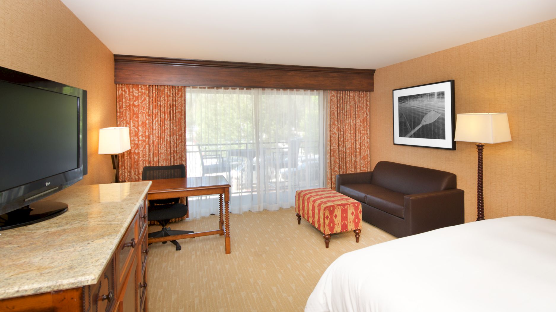 A hotel room with a desk and TV, patterned curtains, a sofa, lamps, and a framed wall print.