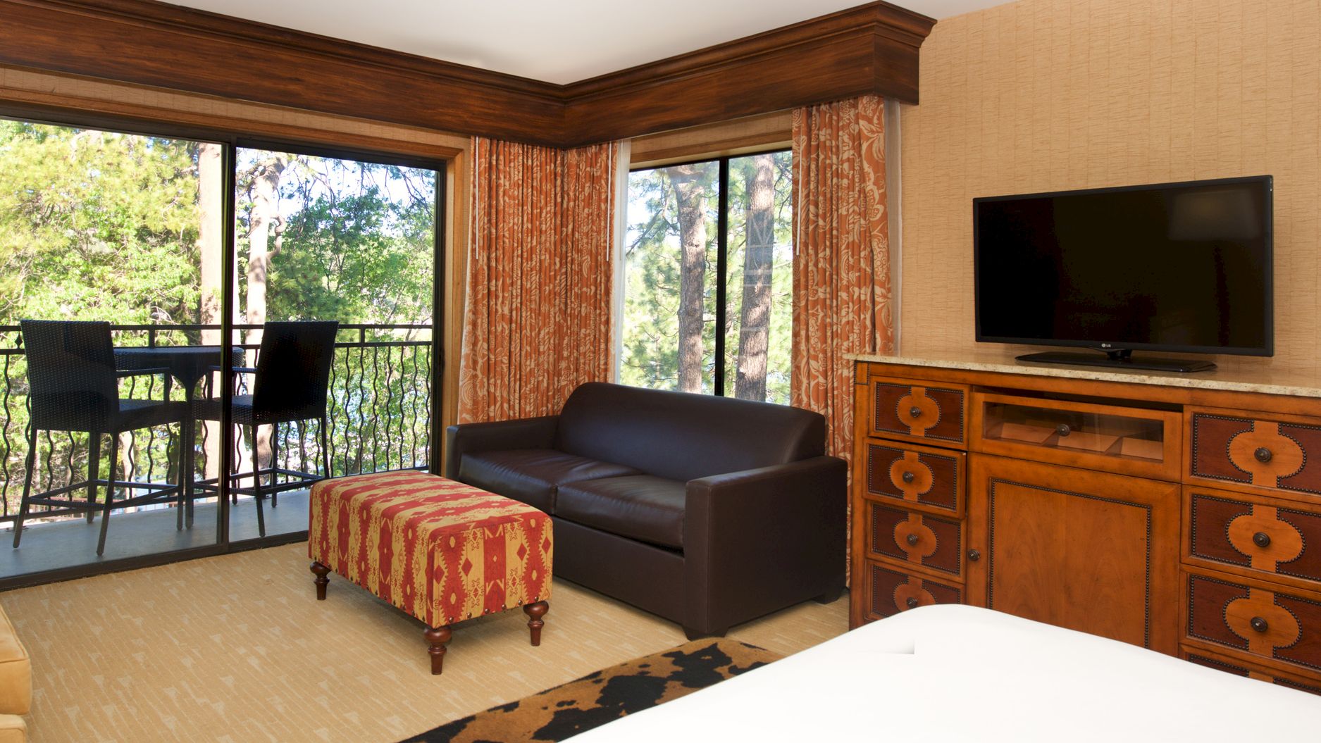Hotel room lounge with large windows, orange curtains, a sofa, and a flat-screen TV on a carved wooden dresser.