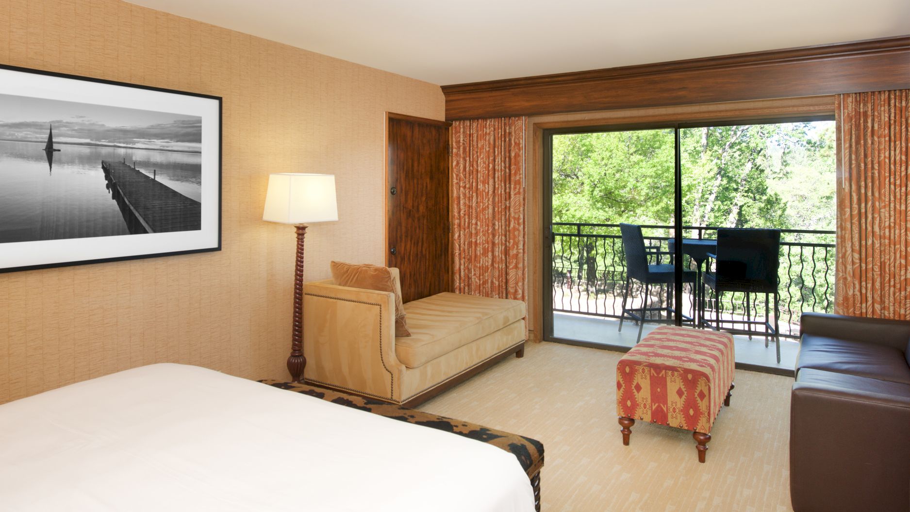 A bright hotel bedroom with a large bed, bedside lamp, patterned ottoman, and large windows/door overlooking green trees.