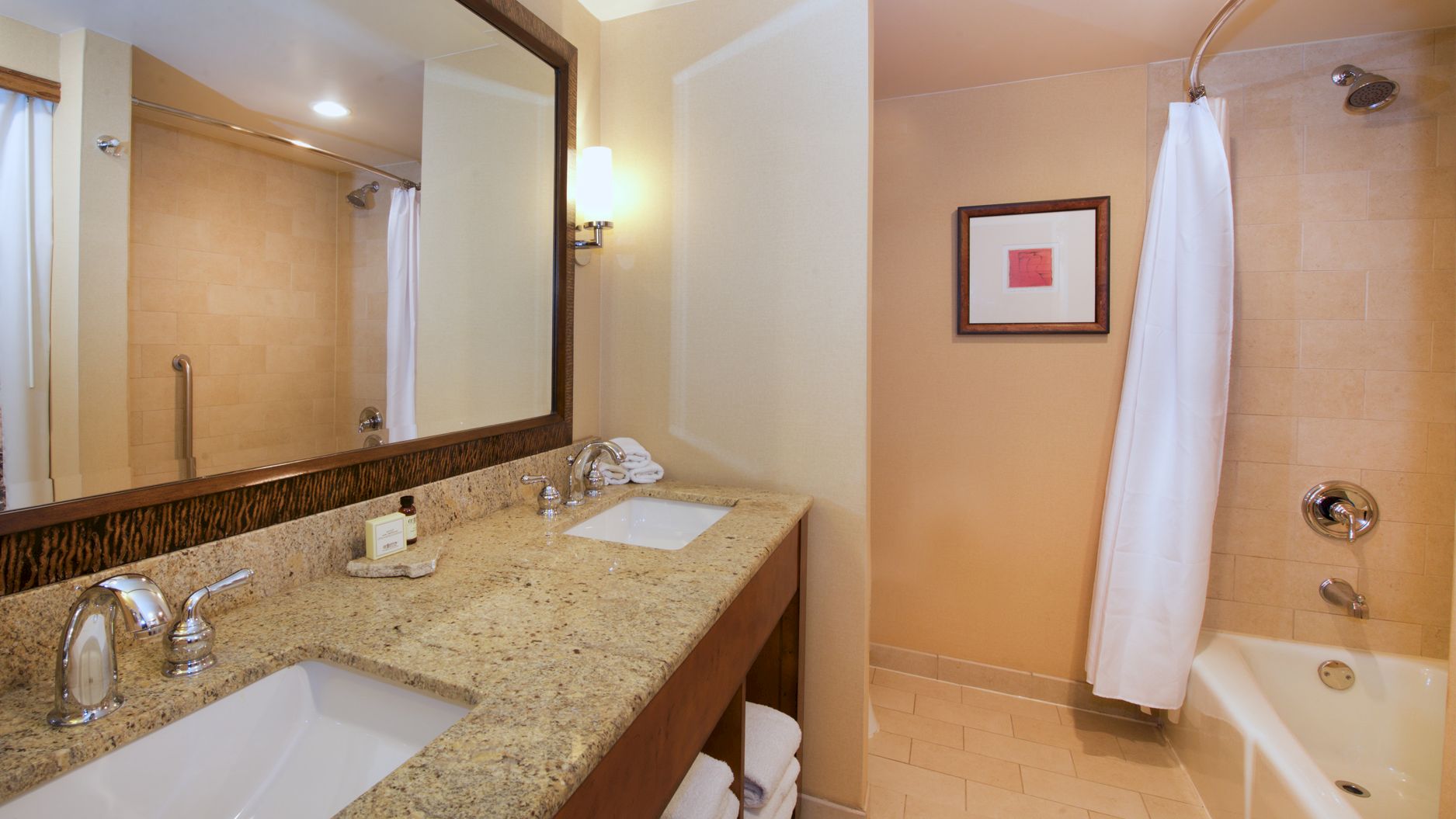 A hotel bathroom with a granite double vanity, large mirror, showerhead, and a bathtub with white towels.