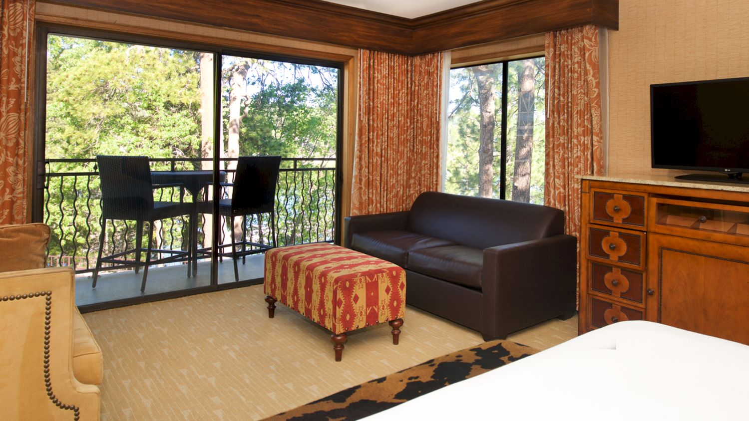 A hotel room with a bed, brown sofa, TV, wooden dresser, patterned curtains, and big windows overlooking green trees.
