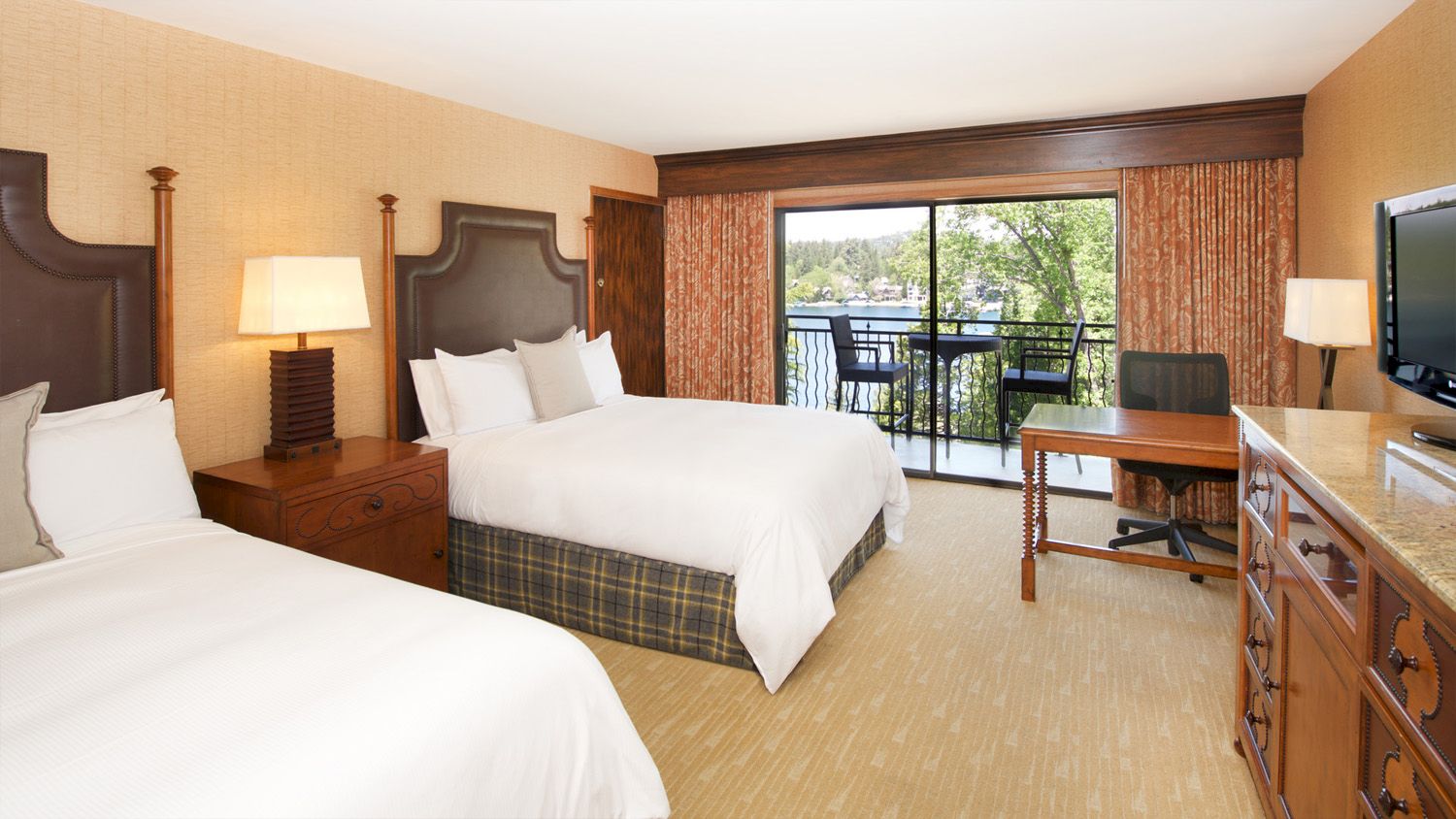 Hotel room with two beds, lamps, a desk with a TV, and big windows opening to a balcony view of trees and water.