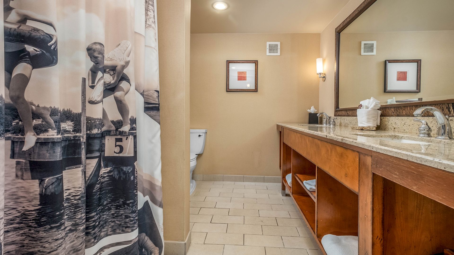 A hotel bathroom with a shower curtain, toilet in the back, and a wooden vanity with sink, mirror, and bright wall lights.