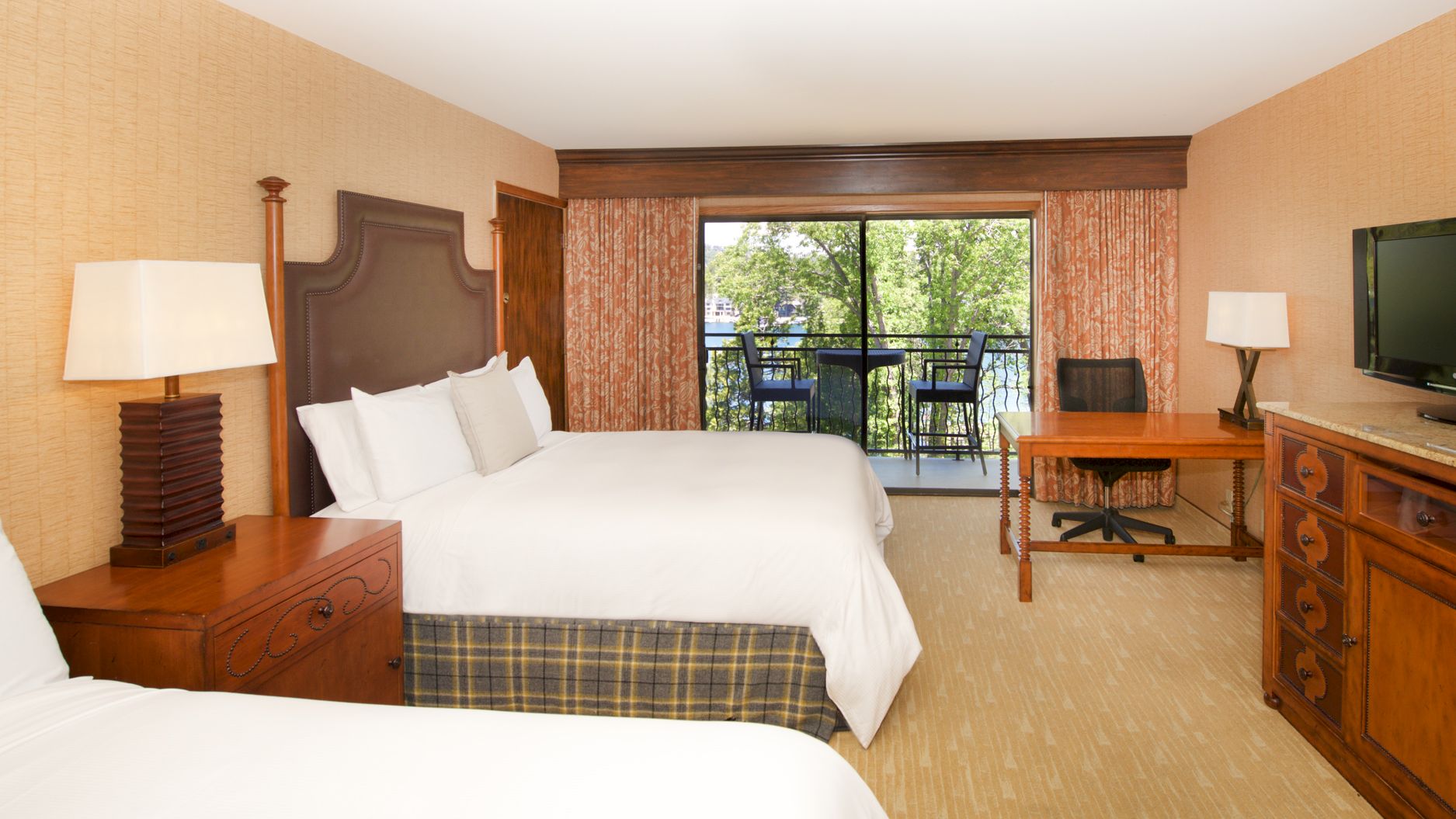 A hotel bedroom with beds, lamps, dresser, flat-screen TV, and patio doors opening to chairs and lush trees outside.