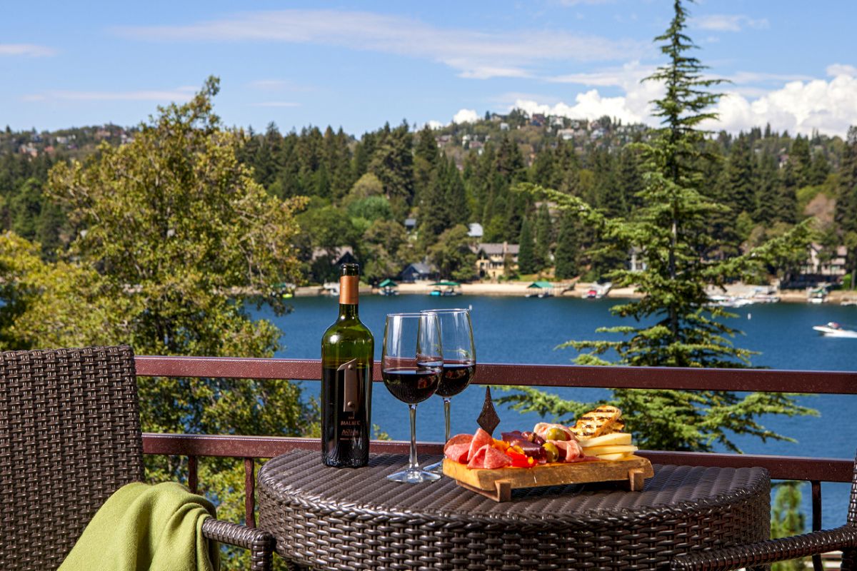 An outdoor table set with a bottle of wine, two glasses, and a cheese board, overlooking a lake, boats, and a pine-lined town.