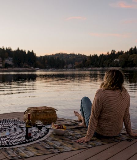A person sits by a calm lake at sunset, surrounded by a picnic spread on a wooden deck.