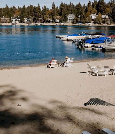 Sunny lakeside with sandy beach, lounge chairs, and a docked row of boats beside a tree-lined town.