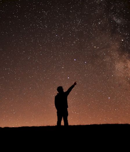 A silhouetted person points into a vast star-filled night sky with the Milky Way glowing across the darkness.