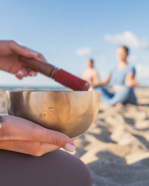A hand holds a metal singing bowl on a sunny beach while another hand gestures, with a couple walking in the background.