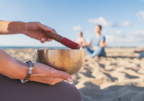 A hand holds a metal singing bowl on a sunny beach while another hand gestures, with a couple walking in the background.