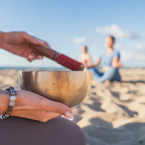 A hand holds a metal singing bowl on a sunny beach while another hand gestures, with a couple walking in the background.