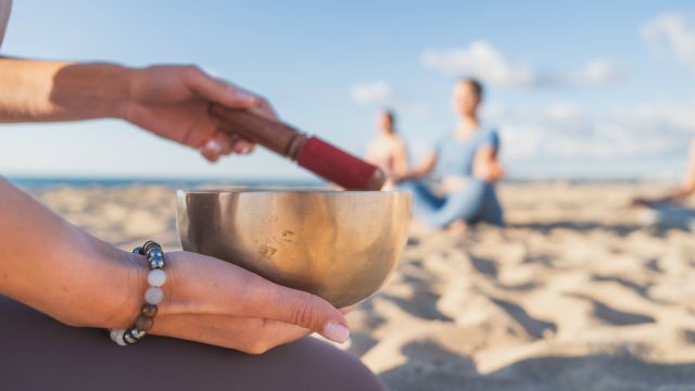 A hand holds a metal singing bowl on a sunny beach while another hand gestures, with a couple walking in the background.