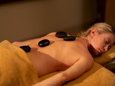 A person lies on a massage table receiving a hot-stone therapy treatment with black stones on their back, relaxing quietly.