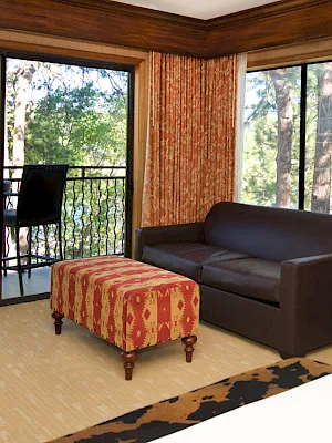 A cozy hotel room with a leather loveseat, bed, dresser with TV, and large windows overlooking lush trees, surrounded by patterned curtains.