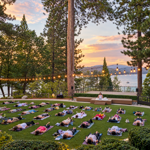 An outdoor yoga class in a green garden at sunset, with people lying on mats beneath string lights.