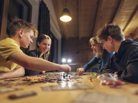 A group of friends sit at a wooden table, smiling and playing a tabletop board game with colorful pieces under warm lights.