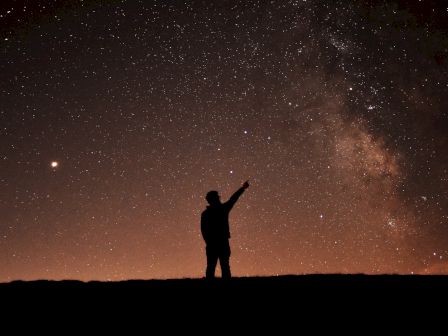 A lone person&rsquo;s silhouette points at a bright, star-filled night sky with the Milky Way visible across the horizon.