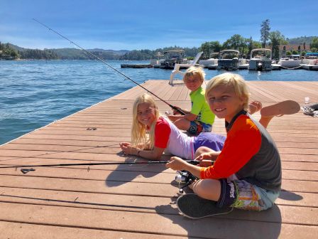 Three kids smile on a dock by a lake, fishing with rods while boats sit in the sunny harbor.