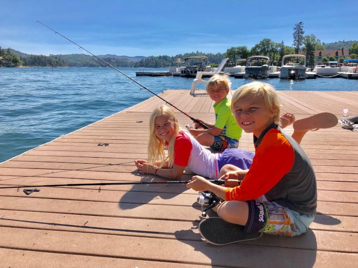 Three kids smile on a dock by a lake, fishing with rods while boats sit in the sunny harbor.