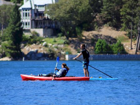 Two people paddle a red ocean kayak on a blue lake while a house and trees line the shore behind them.