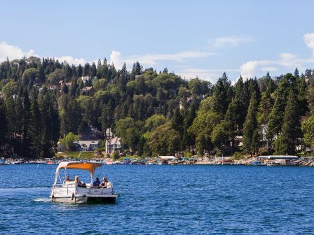 A small boat cruises across a lake toward a marina packed with yachts, with people on board and a forested hillside behind.