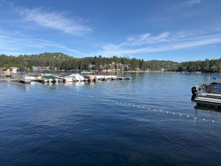 A calm lakeside marina with docked boats, floating platforms, hillside homes, and a blue sky with wispy clouds.