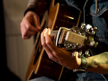 A guitarist in a denim jacket strums an acoustic guitar mid-performance, captured on stage.