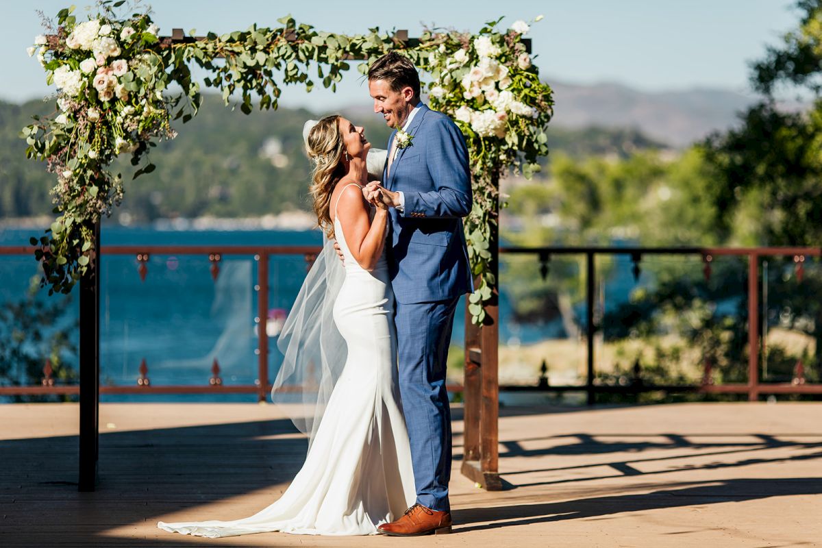 A bride and groom stand under a flower arch by a lakeside, sharing a tender moment at an outdoor wedding ceremony.