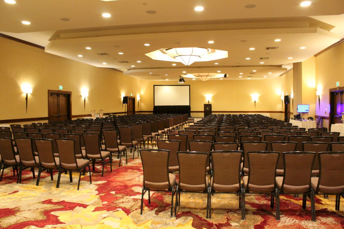 An empty conference room with rows of chairs facing a large screen, projector setup, and bright ceiling lights.