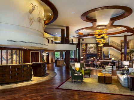 A luxurious lodge-style hotel lobby with a stone fireplace, curved wood beams, chandelier, and mounted deer artwork.