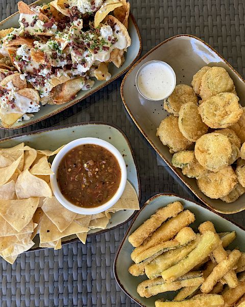 Overhead view of loaded nachos with sour cream, bacon bits, and chives, plus tortilla chips, fries, and dipping sauces.