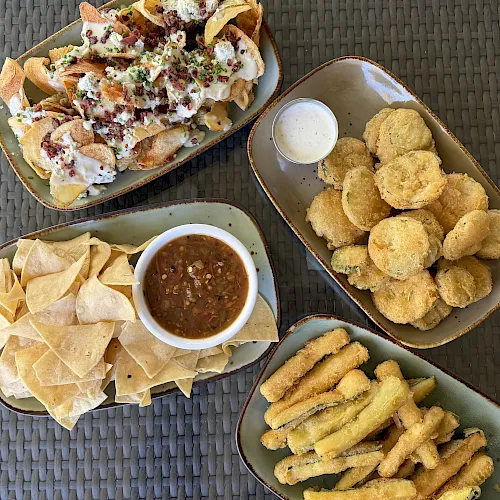 Overhead view of loaded nachos with sour cream, bacon bits, and chives, plus tortilla chips, fries, and dipping sauces.