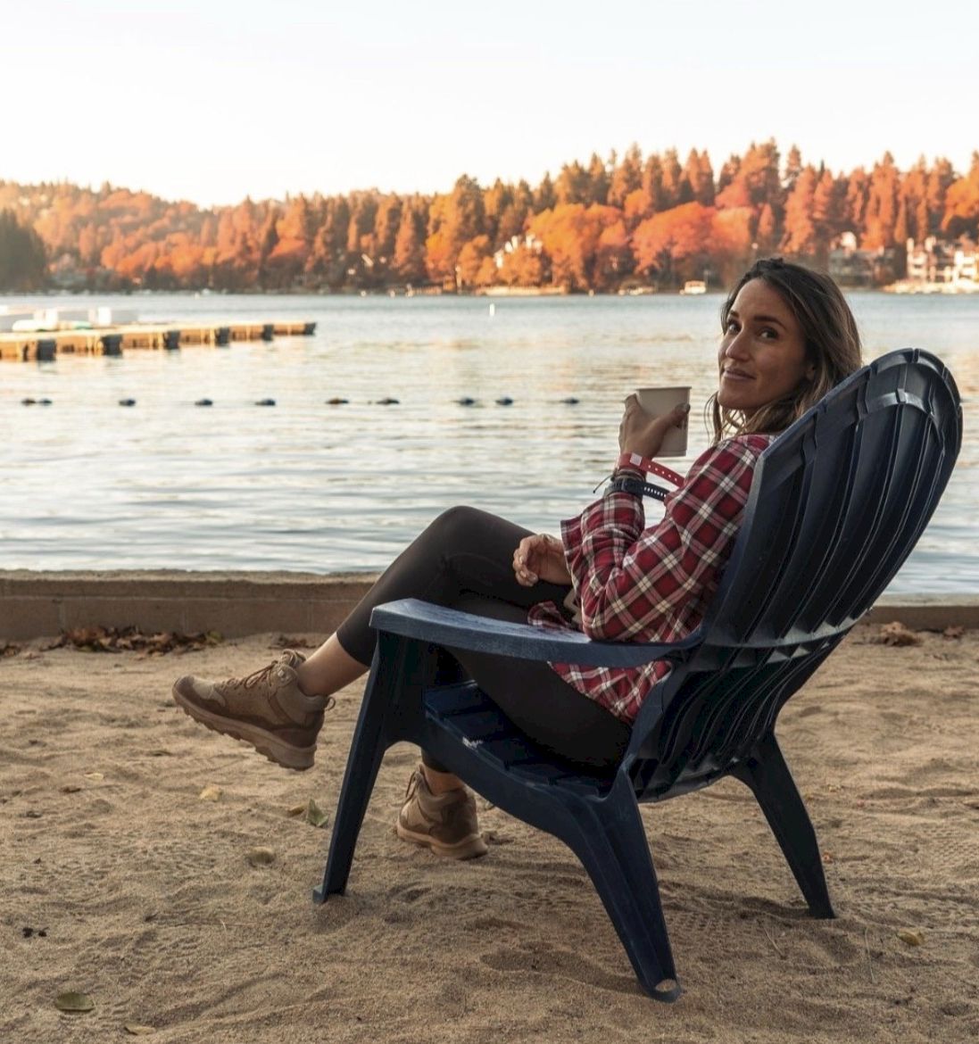 A woman relaxes in a blue chair on a sandy lake shore at sunset, sipping a drink and gazing across the water.