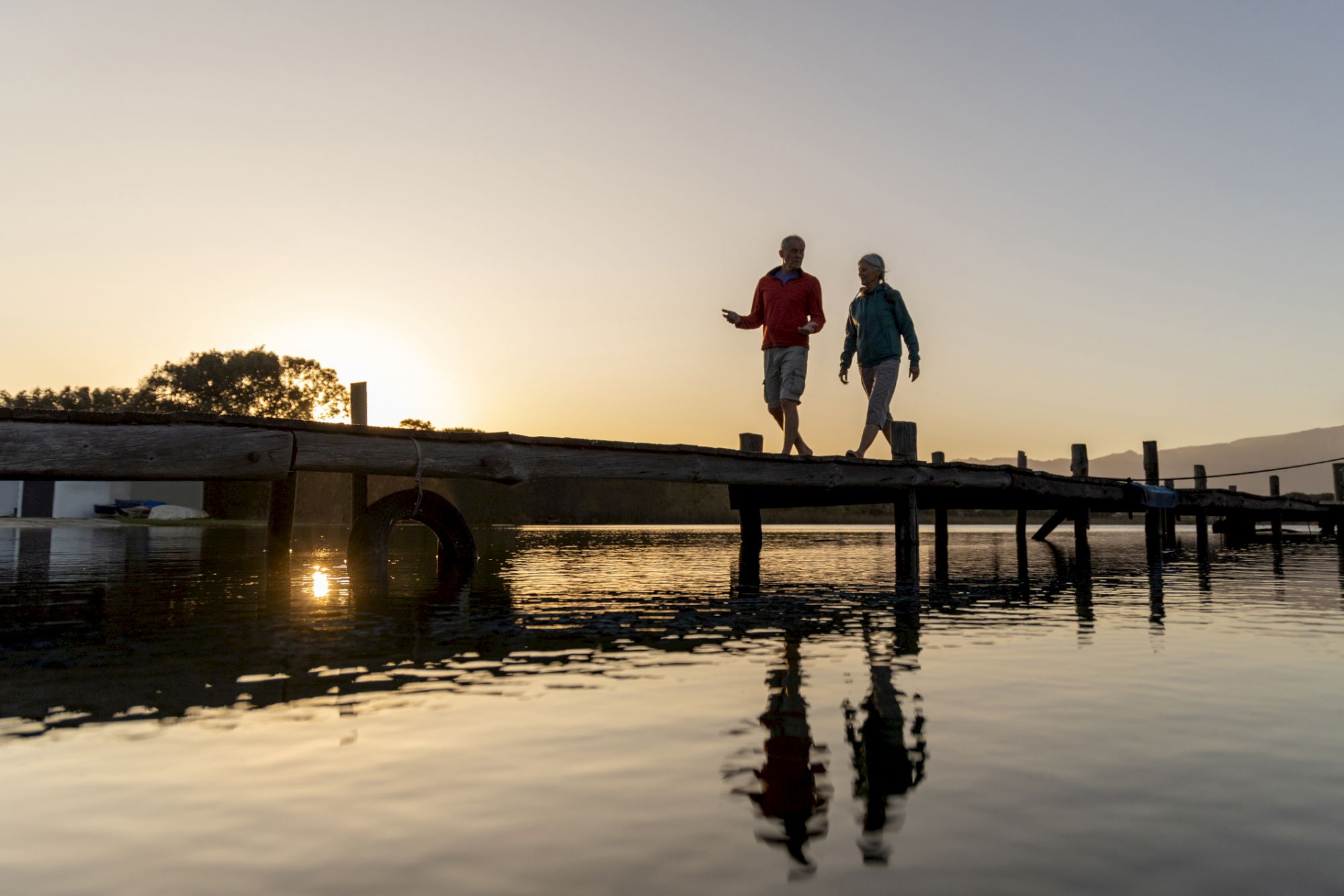 Two people stroll hand-in-hand across a wooden pier at sunset, reflected in the calm water.
