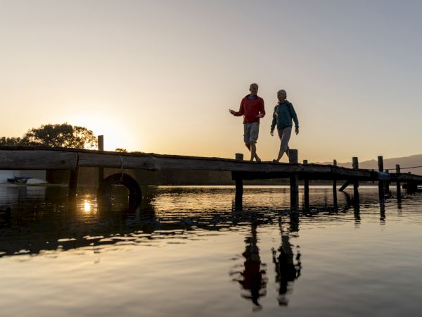 A man and woman stroll on a wooden dock at sunset over calm water, silhouetted against the sky.