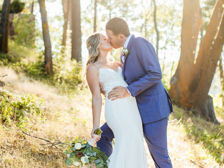 A bride and groom kiss in a sunlit forest, holding a bouquet and wearing wedding attire.