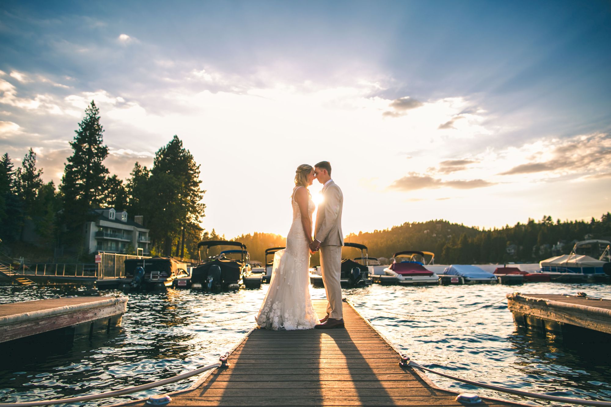 A bride and groom stand on a dock by a lake at sunset, holding hands with boats and cabins nearby.