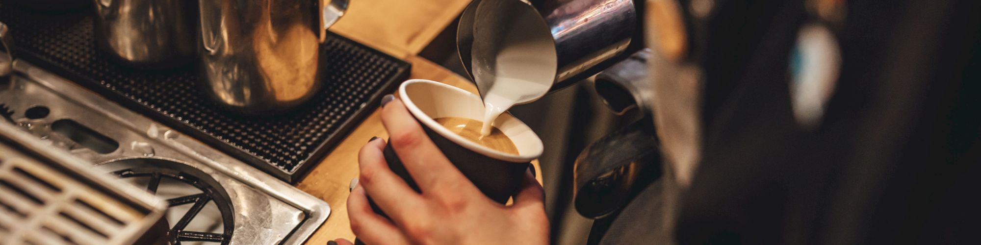 A barista pours steamed milk into a cup of coffee, with a steam wand, espresso machine, and metal pitchers on a wooden counter.
