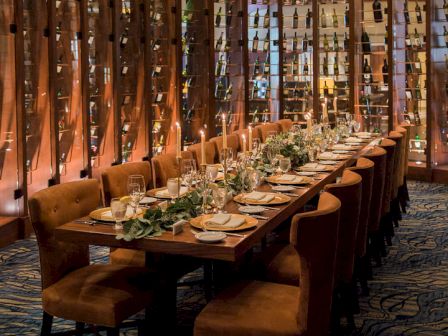 Candlelit restaurant dining room with a long table, chandeliers, and wall-to-wall wine bottle displays.